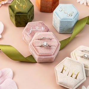 Pode incluir: Uma coleção de caixas hexagonais de veludo para anéis em verde-oliva, laranja queimado, azul-claro, rosa antigo e creme. As caixas apresentam monogramas em folha de ouro ou branca, incluindo N&P, K&B, j&r e F|H. Duas caixas contêm anéis de prata.