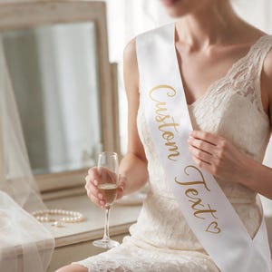 Puede incluir: Una banda de satén blanco con el texto dorado en cursiva Custom Text y un pequeño símbolo de corazón. La banda se lleva en diagonal sobre el torso encima de un vestido de encaje blanco. El material tiene un acabado liso y reflectante.