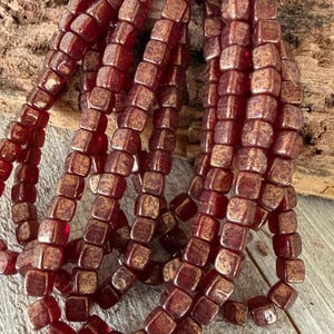 May include: Strands of square, burgundy-colored beads with a metallic gold finish. The beads are strung together, creating a textured and visually appealing pattern. The beads are likely for jewelry making.