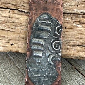 May include: A rectangular copper pendant with a hammered texture and a dark gray, stylized design. The pendant features a central, textured shape with layered details and a decorative swirl. A small hole is at the top for hanging.