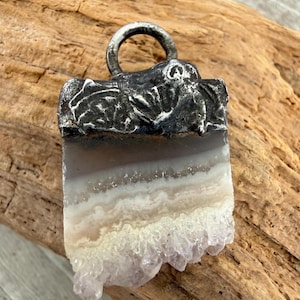 May include: A rectangular pendant featuring a raw agate slice with horizontal bands of grey, white, and translucent beige, ending in a jagged cluster of light purple crystals at the base, topped with a dark, textured metal cap and a circular bail.