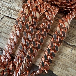 May include: A metallic copper-colored curb chain featuring interlocking oval links with a polished, reflective finish. The chain is arranged in multiple parallel strands, showcasing the uniform shape and smooth texture of the individual metal links.