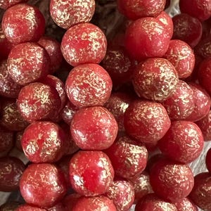 May include: Red glass beads with gold flecks. The beads are round and have a textured surface.