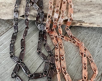 May include: Two metal chains featuring interlocking links shaped like paperclips. One chain has a dark, gunmetal finish, while the other chain has a bright, polished copper finish. Both chains are long and display a repeating geometric pattern.