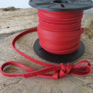 May include: A spool of red leather cord, wound around a black plastic spool. The cord is about 1/4 inch wide and has a smooth, slightly textured surface. A length of the cord is laid out on a wooden surface, tied in a knot.
