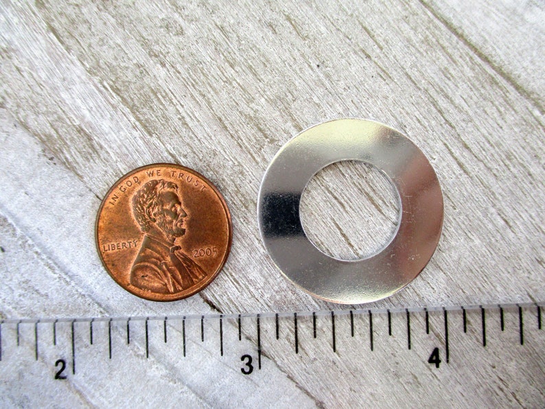 May include: A silver metal washer with a hole in the center, next to a US penny, measured with a ruler. The washer is approximately 1 inch in diameter.