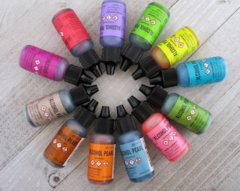May include: A collection of twelve small, colorful alcohol ink bottles arranged in a circular pattern. The bottles have black caps and labels with various colors, including pink, purple, blue, green, and orange. The labels read "Alcohol Pearl".