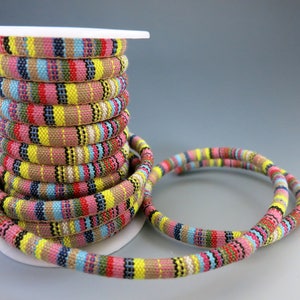 May include: A spool of colorful striped fabric cord. The cord is wound around a white spool and there is a loose loop of the cord in front of the spool. The cord is made of a woven fabric and has a variety of colors, including pink, yellow, blue, and brown.