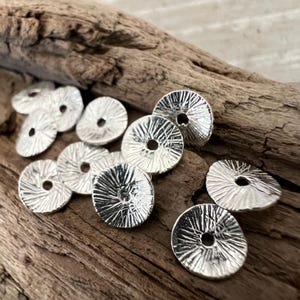 May include: A collection of silver-toned metal beads with a textured, raised design. The beads are round with a hole in the center.