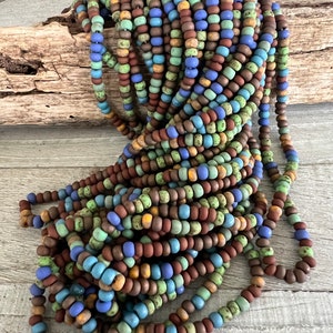 Pode incluir: Uma grande pilha de contas redondas e coloridas em tons de castanho, azul, verde e amarelo. As contas s&atilde;o enfiadas, criando uma apar&ecirc;ncia texturizada e em camadas. As contas est&atilde;o sobre uma superf&iacute;cie de madeira clara.