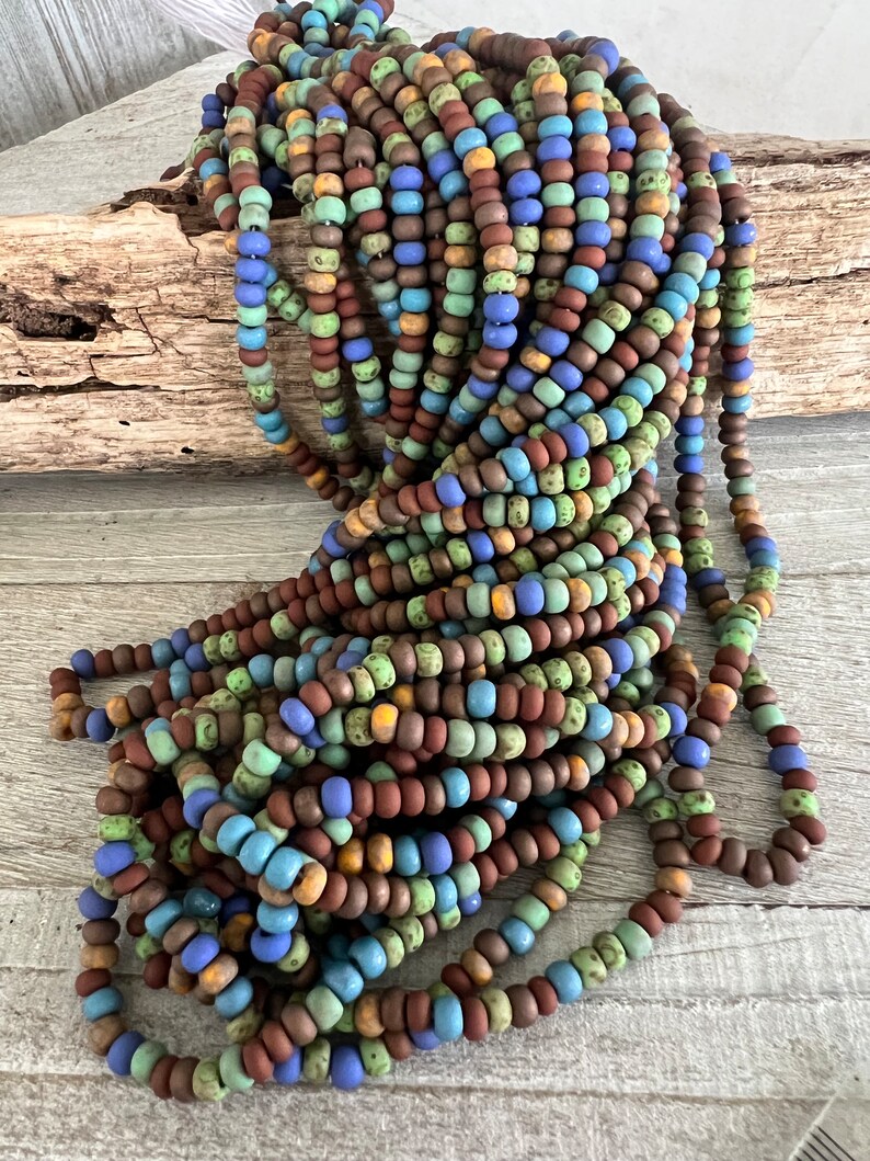 Pode incluir: Uma grande pilha de contas redondas e coloridas em tons de castanho, azul, verde e amarelo. As contas s&atilde;o enfiadas, criando uma apar&ecirc;ncia texturizada e em camadas. As contas est&atilde;o sobre uma superf&iacute;cie de madeira clara.
