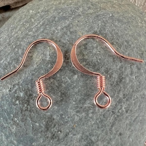 May include: Two copper-coloured earring hooks are displayed against a grey stone background. Each hook has a curved design with a coiled wire detail and a small loop at the bottom for attaching beads or charms. The hooks are designed for jewellery making.