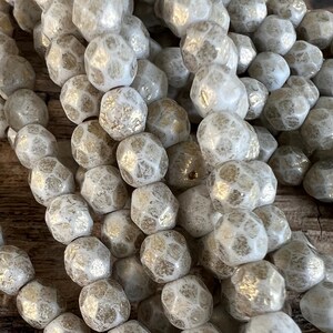 May include: Close-up of a collection of off-white, irregularly shaped beads with a textured surface and gold accents. The beads are clustered together, suggesting they are part of a larger strand or design, suitable for jewelry making.
