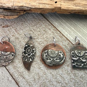 May include: Four metal pendants featuring a combination of copper and silver-toned textured surfaces. Each piece includes a small silver jump ring at the top. The shapes include two circles, one leaf-like form, and one rectangular pendant with rounded corners.