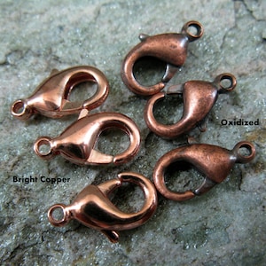 May include: Close-up of several copper-colored lobster claw clasps. Some clasps have a bright copper finish, while others are oxidized. The clasps are designed for jewelry making and crafting. The background is a mottled green and gray surface.
