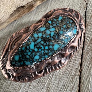 May include: A copper-toned pendant with a turquoise stone inlay. The stone has a mottled pattern of blue and green with flecks of gold.