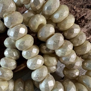 May include: A close-up of a strand of off-white glass beads with a faceted, irregular shape. The beads have a subtle iridescent sheen.