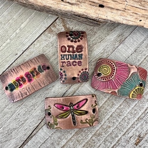 May include: Four ceramic tiles with different designs. One tile has the words "one human race" in brown lettering on a copper background. Another tile has a colorful dragonfly design on a copper background. The third tile has a colorful abstract design with pink, green, and yellow colors. The fourth tile has the word "hope" in rainbow colors on a copper background.