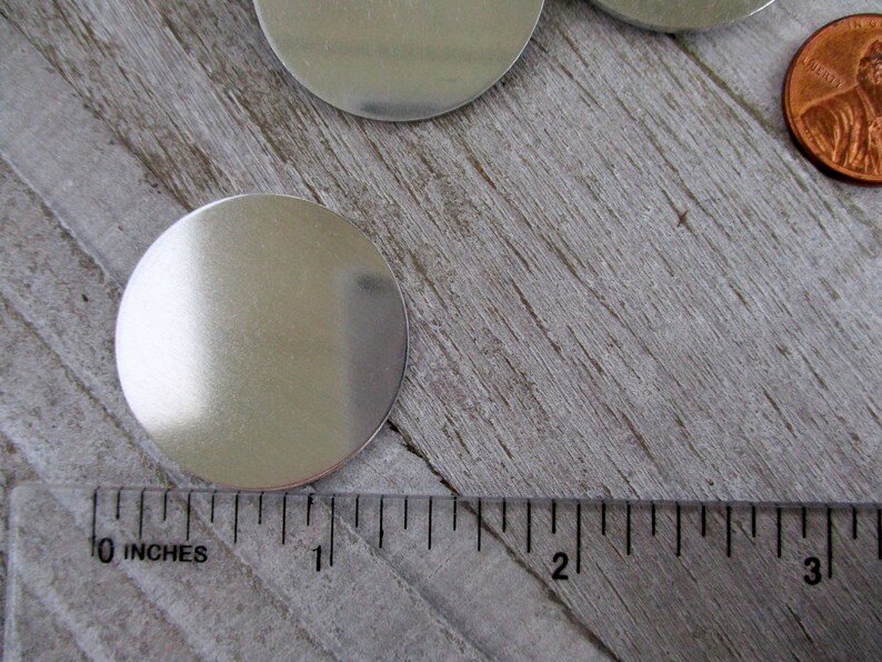 May include: A silver metal disc measuring 1 inch in diameter. The disc is smooth and reflective, and it is sitting on a wooden surface.