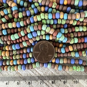 Pode incluir: Uma cole&ccedil;&atilde;o de contas coloridas, com acabamento fosco, em tons de azul, verde, marrom e amarelo. As contas s&atilde;o cil&iacute;ndricas e enfiadas. Uma moeda de um centavo &eacute; colocada ao lado das contas para refer&ecirc;ncia de tamanho. Uma r&eacute;gua est&aacute; na parte inferior.