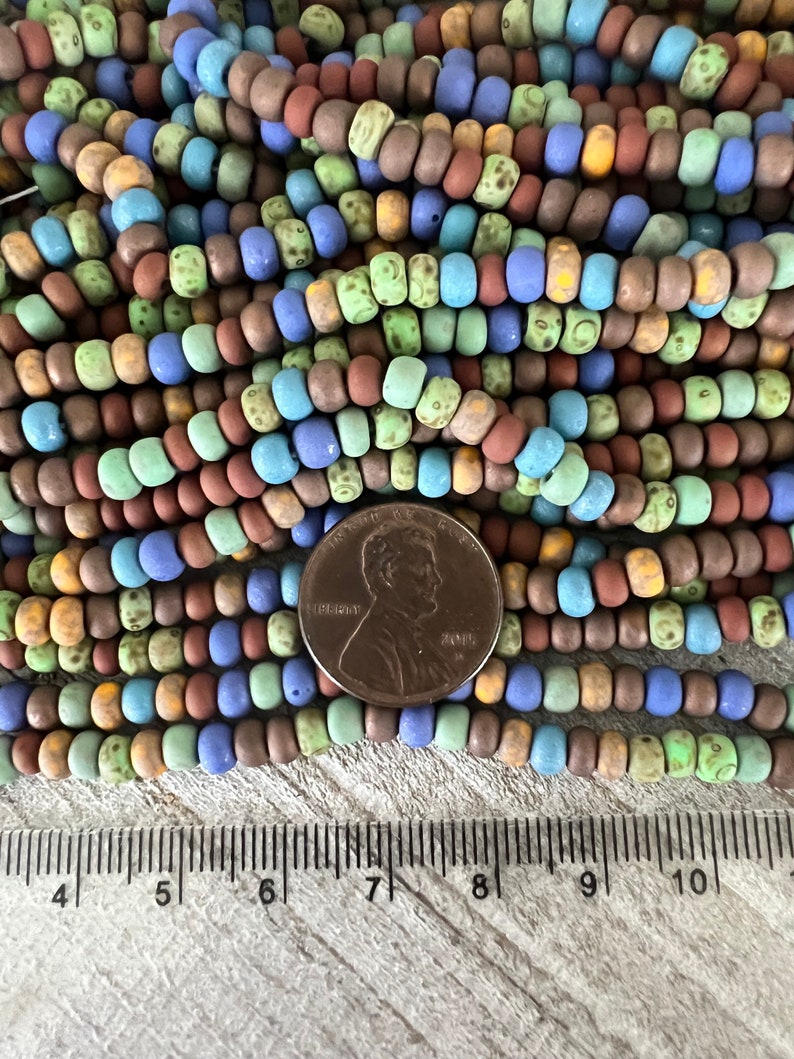 Pode incluir: Uma cole&ccedil;&atilde;o de contas coloridas, com acabamento fosco, em tons de azul, verde, marrom e amarelo. As contas s&atilde;o cil&iacute;ndricas e enfiadas. Uma moeda de um centavo &eacute; colocada ao lado das contas para refer&ecirc;ncia de tamanho. Uma r&eacute;gua est&aacute; na parte inferior.