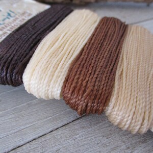 May include: Three spools of waxed cord in brown, cream, and tan colors. The spools are labeled "Knot-it!" and "Waxed Brazilian Cordon Baguá  Cordón Baguá".