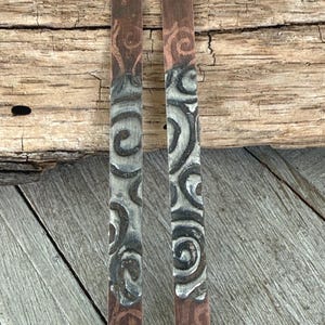 May include: Two rectangular copper earrings with a decorative swirl design in silver and black. The top and bottom sections of the earrings have a reddish-brown patina. The earrings are displayed on a wooden surface.