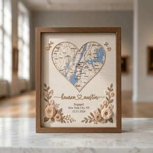 May include: A framed wall art piece featuring a heart-shaped map of New York City. The design includes embroidered floral accents in beige and cream tones, the names "lauren" and "austin" with a heart, and the text "Engaged New York City, NY 12.31.2024".