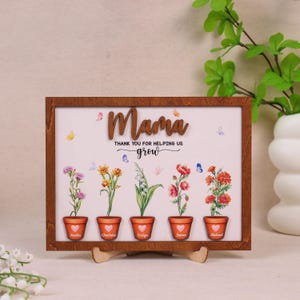 May include: A wooden framed sign with the word "Mama" in raised letters. The sign features five flower pots with names, and the text "THANK YOU FOR HELPING US grow". The flowers are colorful, and butterflies are scattered around the design.