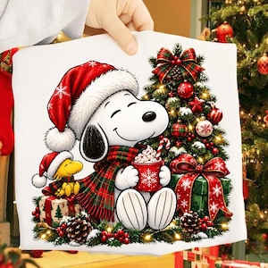 May include: A white t-shirt featuring a graphic of Snoopy wearing a red Santa hat and a plaid scarf, holding a red mug with a snowflake design. Beside Snoopy is Woodstock in a Santa hat, a decorated Christmas tree, and wrapped gift boxes.
