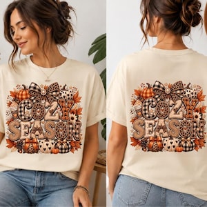 Könnte beinhalten: Ein cremefarbenes Kurzarm-T-Shirt mit einem Grafikdruck und dem Text COZY SEASON. Das Design zeigt Kürbisse, Herbstblätter, eine große Schleife sowie verschiedene Karomuster und Leopardenmuster in den Farben Orange, Braun und Schwarz.