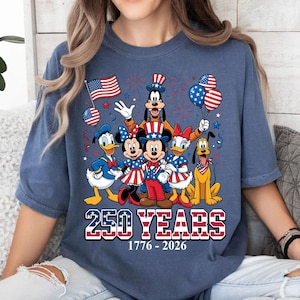 May include: Blue t-shirt featuring a graphic of Disney characters dressed in patriotic attire, celebrating 250 years of American history. The design includes Mickey and Minnie Mouse, Donald Duck, Goofy, Daisy Duck, and Pluto, with fireworks and balloons.