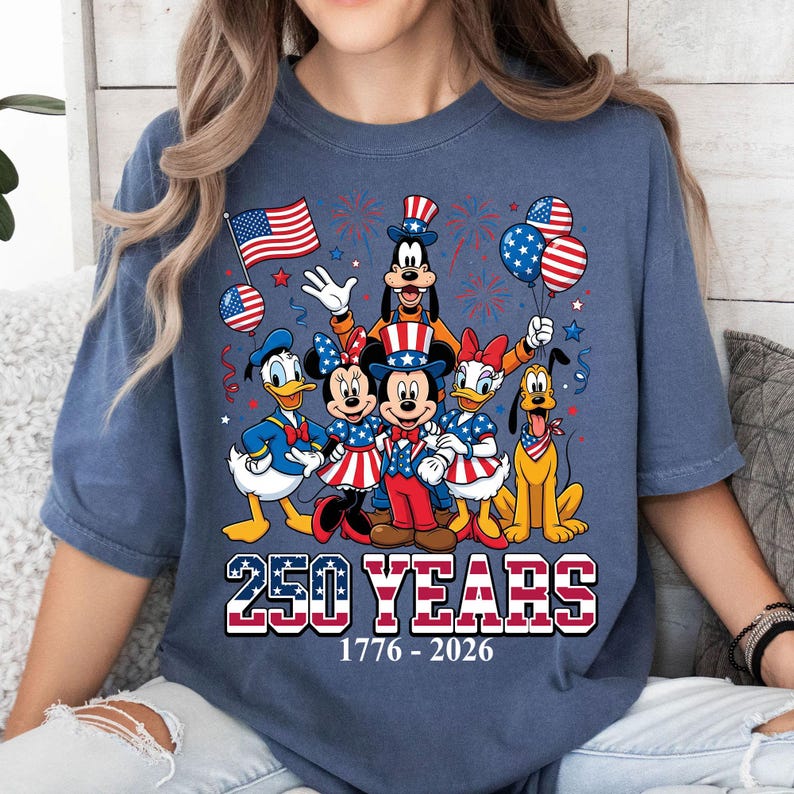 May include: Blue t-shirt featuring a graphic of Disney characters dressed in patriotic attire, celebrating 250 years of American history. The design includes Mickey and Minnie Mouse, Donald Duck, Goofy, Daisy Duck, and Pluto, with fireworks and balloons.