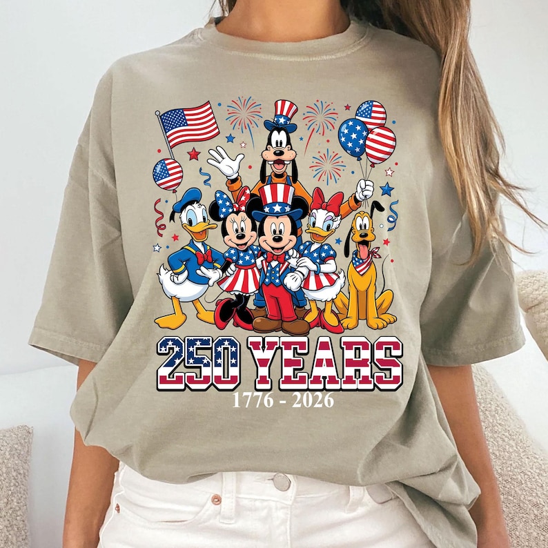 May include: A beige t-shirt featuring a graphic of Disney characters Mickey, Minnie, Donald, Daisy, Goofy, and Pluto, dressed in patriotic attire. The design includes fireworks, balloons, and an American flag, with the text "250 YEARS 1776-2026".