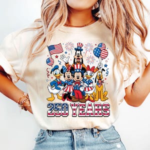 May include: A cream-colored t-shirt featuring a graphic of Disney characters dressed in patriotic attire, including Mickey and Minnie Mouse, Donald Duck, and Goofy. The design includes an American flag, fireworks, and the text "250 YEARS".