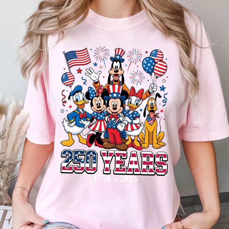 May include: Pink t-shirt featuring a graphic of Disney characters Mickey Mouse, Minnie Mouse, Donald Duck, Daisy Duck, Goofy, and Pluto, dressed in patriotic attire. The shirt includes the text "250 YEARS" and the dates "1776-2026".