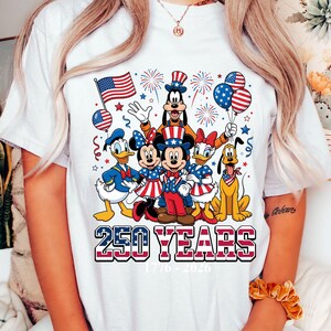 May include: White t-shirt featuring a colorful graphic of Disney characters dressed in patriotic attire. The design includes Mickey and Minnie Mouse, Donald and Daisy Duck, Goofy, and Pluto, with an American flag and balloons. The text "250 YEARS 1776-2026" is also displayed.