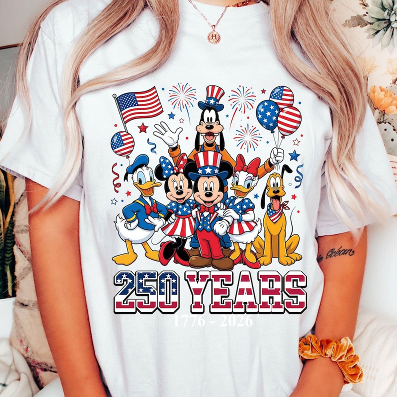 May include: White t-shirt featuring a colorful graphic of Disney characters dressed in patriotic attire. The design includes Mickey and Minnie Mouse, Donald and Daisy Duck, Goofy, and Pluto, with an American flag and balloons. The text "250 YEARS 1776-2026" is also displayed.