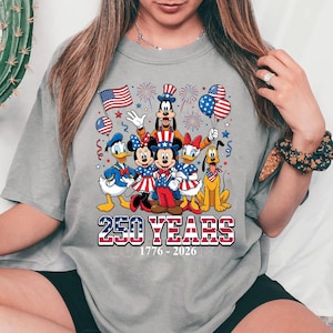 May include: Gray t-shirt featuring a patriotic design with Disney characters. Mickey, Minnie, Donald, Daisy, Goofy, and Pluto are dressed in red, white, and blue. The text "250 YEARS 1776-2026" is displayed below the characters.