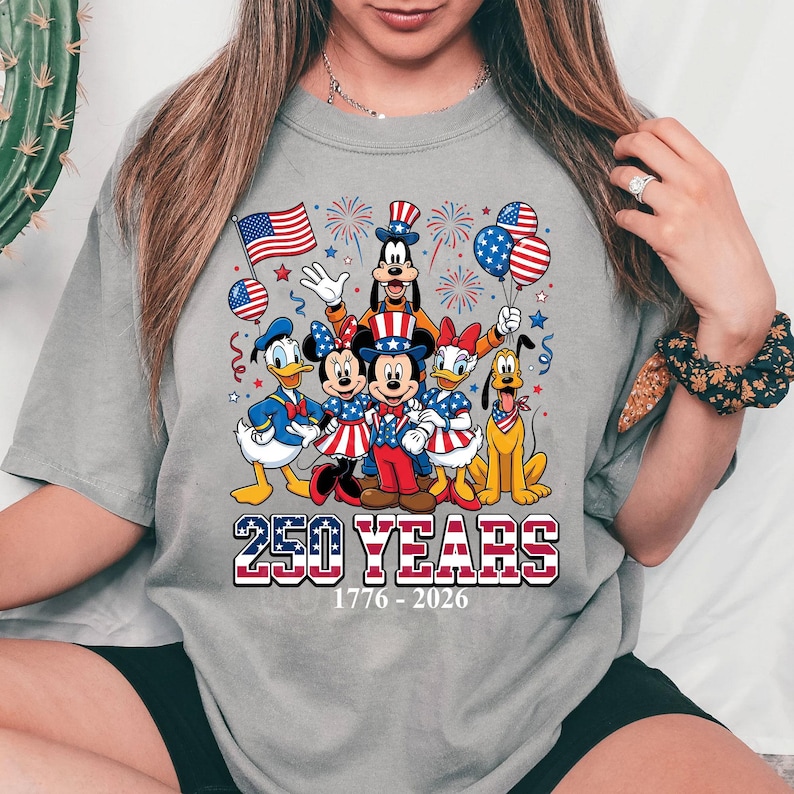 May include: Gray t-shirt featuring a patriotic design with Disney characters. Mickey, Minnie, Donald, Daisy, Goofy, and Pluto are dressed in red, white, and blue. The text "250 YEARS 1776-2026" is displayed below the characters.
