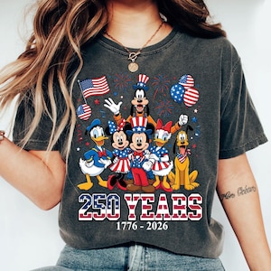 May include: Dark gray t-shirt featuring a graphic of Disney characters in patriotic attire. The design includes Mickey and Minnie Mouse, Donald and Daisy Duck, Goofy, and Pluto, with American flags and fireworks. Text reads "250 YEARS 1776-2026".