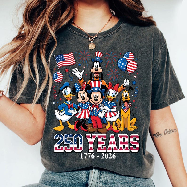 May include: Dark gray t-shirt featuring a graphic of Disney characters in patriotic attire. The design includes Mickey and Minnie Mouse, Donald and Daisy Duck, Goofy, and Pluto, with American flags and fireworks. Text reads "250 YEARS 1776-2026".