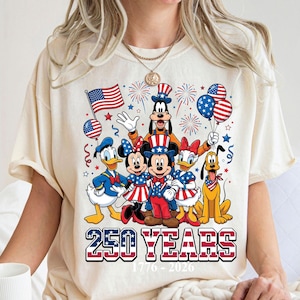 May include: Cream-colored t-shirt featuring a patriotic design with Disney characters. Mickey, Minnie, Donald, Daisy, Goofy, and Pluto are dressed in red, white, and blue. The shirt includes the text "250 YEARS 1776-2026" in a patriotic font.