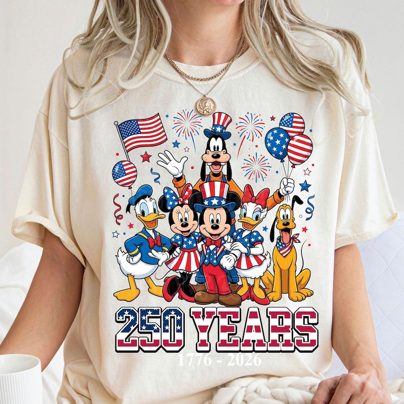 May include: Cream-colored t-shirt featuring a patriotic design with Disney characters. Mickey, Minnie, Donald, Daisy, Goofy, and Pluto are dressed in red, white, and blue. The shirt includes the text "250 YEARS 1776-2026" in a patriotic font.