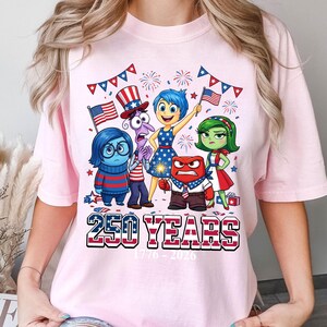 Può includere: T-shirt rosa chiaro con una grafica di personaggi animati e motivi della bandiera americana. Il testo "250 YEARS 1776-2026" è visualizzato in rosso, bianco e blu. Il design include fuochi d'artificio ed elementi patriottici.