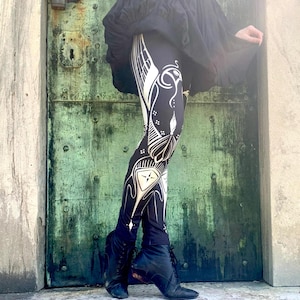 May include: Black leggings with a white Art Nouveau-inspired design. The leggings feature a repeating pattern of stylized floral and geometric shapes. The person is wearing black boots and a black dress. The background is a weathered green door.