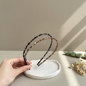 Op de afbeelding: Twee haarbanden met een geweven ontwerp. Eén is zwart, de andere bruin met een patroon. Ze worden gepresenteerd op een wit, rond, getextureerd oppervlak. De achtergrond is neutraal van kleur.