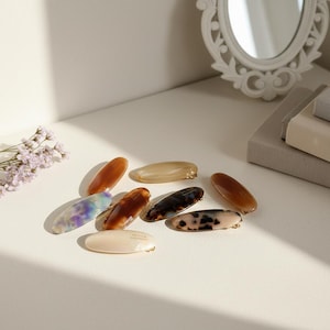 May include: A collection of oval-shaped hair clips in various colors and patterns, including tortoiseshell, marbled, and solid hues. The clips are arranged on a white surface, with a decorative mirror and books in the background.