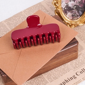 Puede incluir: Una pinza para el pelo de color burdeos brillante. Tiene forma rectangular con la parte superior curvada y dientes para sujetar el cabello. El accesorio para el cabello está hecho de un material liso y brillante, de unos 10 cm.