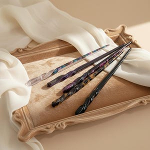 Puede incluir: Cinco palillos para el pelo de varios colores y diseños descansan sobre una bandeja decorativa. Los palillos incluyen diseños transparentes, morados, negros y carey. La bandeja es de color marrón claro con un borde decorativo, y un paño blanco está drapeado en el fondo.
